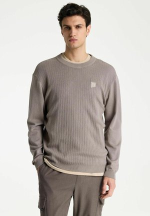 Mann trägt einen taupefarbenen, gerippten Pullover mit kleinem Logo auf der Brust und passende taupefarbene Cargohosen vor einem schlichten hellen Hintergrund.
