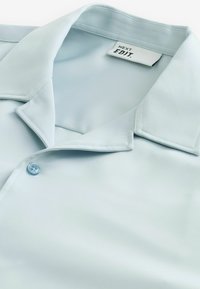 Camisa de manga corta de color azul claro con una textura suave y brillante, un collar puntiagudo y un cierre de un solo botón. La etiqueta dice "NEXT EDIT."