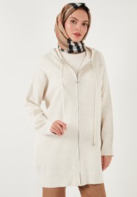 Bežová mikina na zips s kapucňou a šnúrkami. Mäkkučká, textúrovaná látka s dlhými rukávmi, doplnená vzorovaným šatkou.