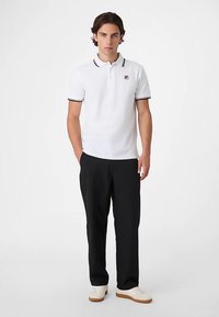 Valkoinen poolopaita, jossa on kontrastiväriset mustat reunukset ja pieni logo vasemmassa rinnassa. Yhdistetty mustiin housuihin ja vaaleisiin kenkiin.