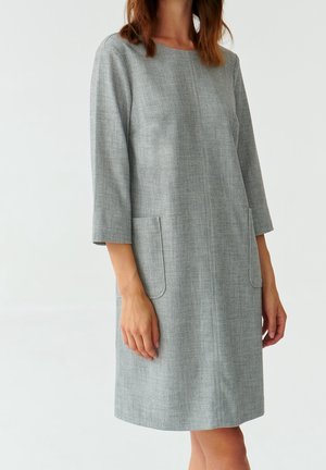 Frau trägt ein hellgraues, knielanges Kleid mit dreiviertel Ärmeln und Fronttaschen, steht vor einem einfachen weißen Hintergrund.