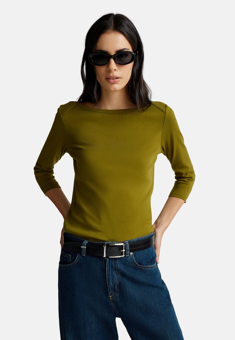 Mujer con cabello negro que lleva gafas de sol negras, una blusa verde oliva de manga tres cuartos, jeans azules y un cinturón negro con hebilla plateada, con las manos detrás de la espalda.