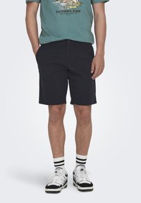 Shorts noirs avec un devant plat, deux poches latérales et une fermeture à bouton, associés à un T-shirt graphique vert et des baskets blanches avec des accents noirs.