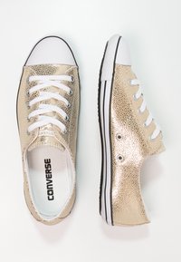 Zapatillas de lona texturizadas doradas con cordones blancos, suela de goma negra y puntera blanca. Logo de Converse visible en la plantilla.