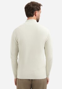 Man in een lichtbeige geribde lange mouwen sweater, met zijn rug naar de camera, gecombineerd met een bruine broek, tegen een effen achtergrond.