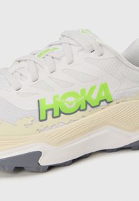Bijela sportska cipela s mrežastim gornjim dijelom, zelenim i crnim "HOKA" logotipom, te teksturiranim kremastim međuslojem. Ima sivi gumeni potplat.