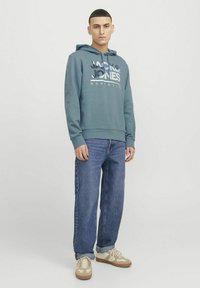 Hoodie azul grisáceo con texto impreso en la parte frontal, puños elásticos y cintura ajustada, combinado con jeans de mezclilla azul claro y zapatillas beige.