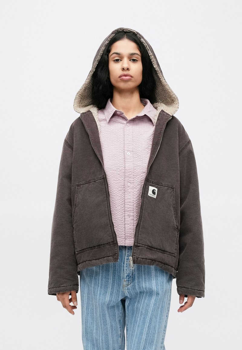 Dark gray hooded jacket with a cream, fuzzy lining, featuring front pockets and a small logo patch, worn over a light pink textured shirt.