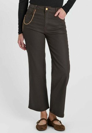 Vrouw draagt donkerbruine wijde broek met gouden kettingdetail, zwart langarmtopje en bruine platte schoenen met gespsluitingen.