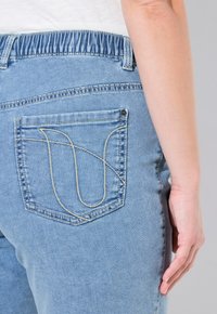 Jeans de mezclilla azul claro con una cinturilla elástica, que presentan un bolsillo trasero con un patrón de costura beige curvado y un detalle de remache plateado.