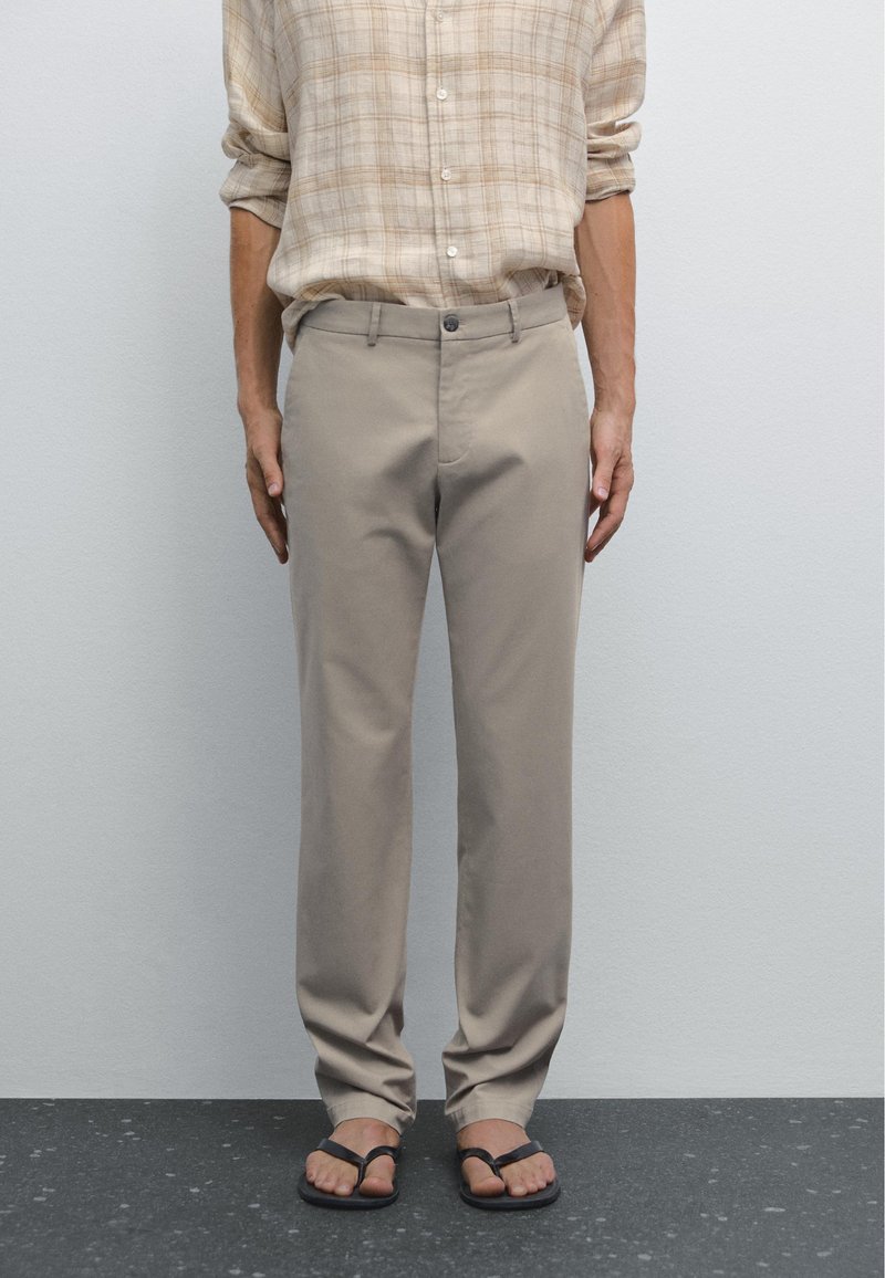 Beige chinos with a straight fit, made of smooth cotton fabric, featuring a button closure and pockets, worn with black flip-flops.