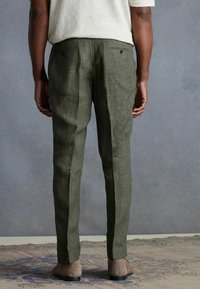 Olive green linen trousers with two back pockets and subtle texture. The fit is tapered, complementing gray suede shoes.