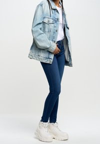 Chaqueta de denim azul claro sobre una camiseta blanca, combinada con jeans skinny azul oscuro y zapatillas blancas gruesas. Atuendo simple y casual.