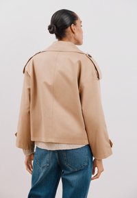 Tan gekropte jas met een iets oversized pasvorm, voorzien van een brede kraag, schouderknopen en gladde stof. Gedragen over een lichte trui en jeans.