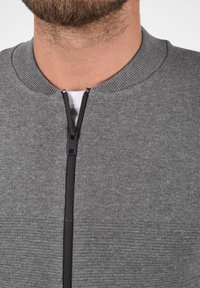 Siva zip-up jopica z rebrastim ovratnikom in teksturirano blago. Ima vidno črno zadrgo spredaj, z belo notranjo plastjo, ki se kaže.