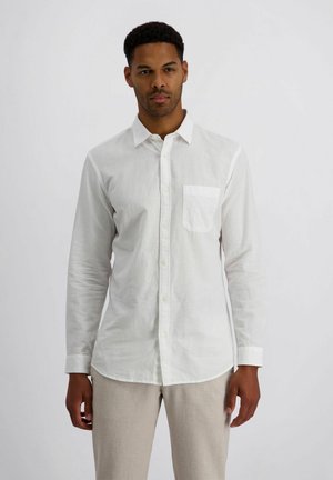 Man staat, draagt een wit overhemd met lange mouwen en een borstzak en beige broek, tegen een effen lichte achtergrond.