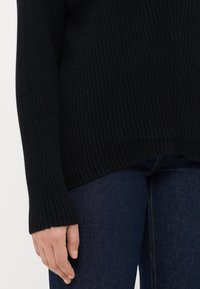 Person som bär en svart ribbad tröja med långa ärmar och mörkblå denimjeans, handen avslappnad vid sidan.