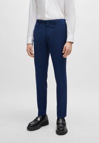 Marineblaues, maßgeschneidertes Hosenpaar mit Gürtelschlaufen und Fronttaschen, aus glattem Stoff gefertigt. Getragen mit einem weißen Hemd und schwarzen Schuhen.