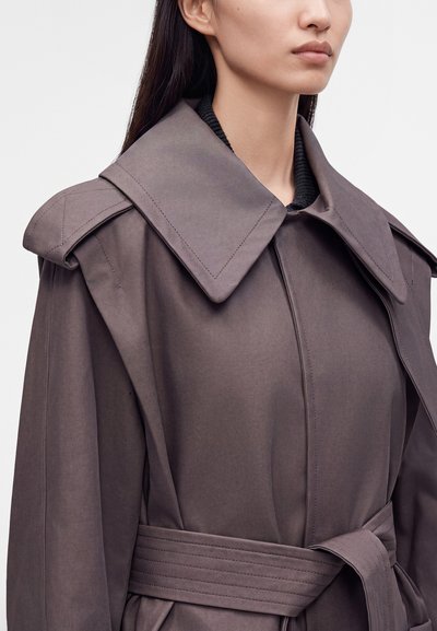 Γυναίκα που φορά ένα μουτζουρωμένο μωβ oversized παλτό με φαρδύ γιακά και ζώνη στη μέση, εμφανίζεται από τους ώμους έως το μέσο του κορμού.