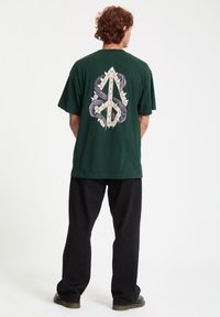 Grøn t-shirt med korte ærmer, der har et stort grafik af sammenflettede slanger og pigge i lilla og beige på ryggen.