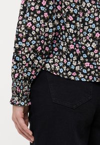 Svart blomstret bluse med rosa, blå og hvite blomster. Har elastiske mansjetter og en avslappet passform, kombinert med mørke bukser.