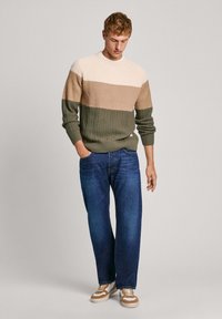 Suéter de punto en colores crema, beige y verde oliva con rayas, que presenta un patrón texturizado; combinado con jeans azules y zapatillas marrones.