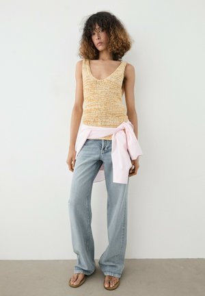 Jeune femme aux cheveux bouclés portant un haut en tricot beige sans manches, un jean ample bleu clair, une chemise rose nouée à la taille et des sandales marron.
