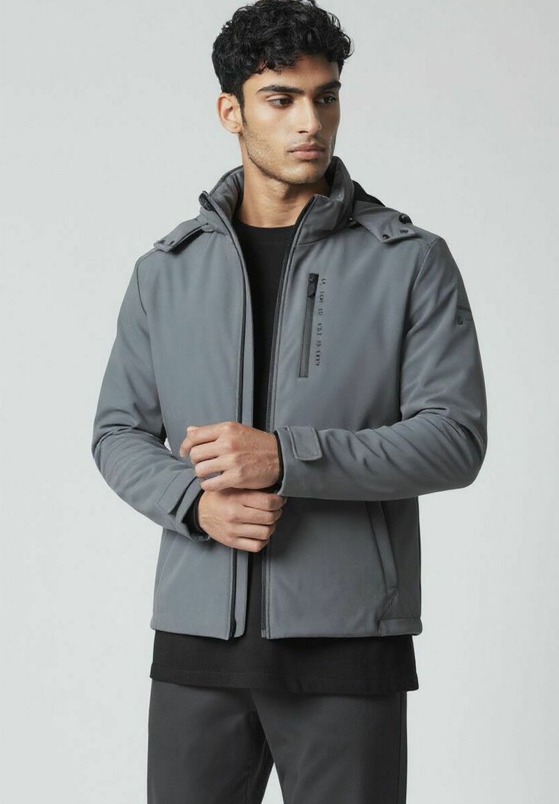 Veste softshell grise avec un front zippé, capuche ajustable et accents à boutons, portée sur une chemise noire. Présente un design épuré et une texture lisse.