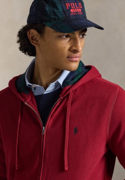 Sweatshirt com capuz vermelho e fecho éclair, com cordões, sobreposto a um suéter azul-marinho e uma camisa azul clara com gola. Inclui um boné xadrez escuro com logótipo.