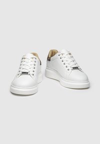Zapatillas blancas hechas de cuero suave, con punta redondeada, cierre de cordones en la parte delantera y un sutil acento marrón en el talón. Suela blanca gruesa.