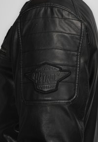 Schwarze Lederjacke mit gesteppten Schulterdetails und einem strukturierten Patch mit dem "Petrol"-Logo. Glatte Oberfläche mit sichtbaren Nähten.