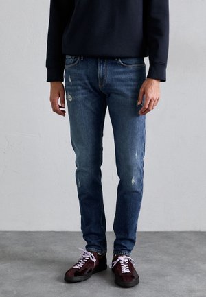 Mann trägt Slim-Fit-blaue Jeans mit leichten Gebrauchsspuren, schwarzen Sweatshirt und dunkelrote Sneaker mit weißen Schnürsenkeln und steht auf grauem Boden.