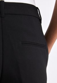 Prim plan cu o persoană care poartă pantaloni negri croiți pe măsură, cu buzunar lateral și buclă pentru curea, asortați cu un top alb texturat.