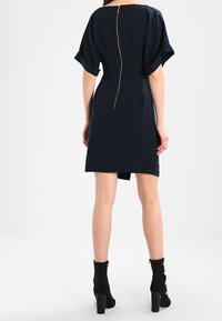 Granatowa krótka sukienka z bufiastymi rękawami, z zamkiem błyskawicznym z tyłu, dopasowanym stanem i prostą spódnicą. Noszona z czarnymi botkami na obcasie.