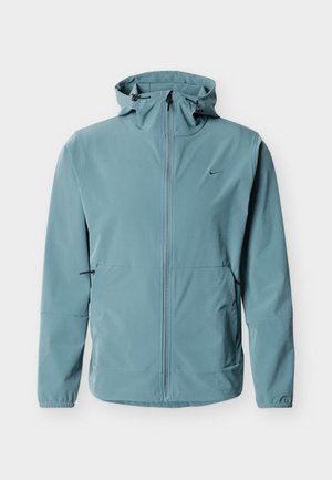 Vaaleansininen hupullinen takki, jossa on täysi etuvetoketju, joustavat hihansuut, sivutaskut ja pieni musta Nike-logo vasemmalla rinnalla.