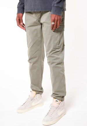 Pantalon cargo gris clair avec poches latérales et manches élastiques, associé à des baskets montantes beiges arborant un logo Nike blanc.
