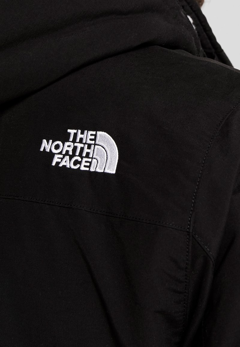 Veste noire avec logo "The North Face" brodé en blanc sur la partie supérieure du dos, près de la couture de l'épaule.