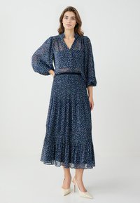 Navy jurk met bloemenpatroon, voorzien van een v-hals, ballonmouwen en een gelaagde rok. Gemaakt van lichte stof met een vloeiende textuur.