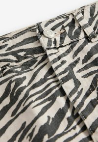 Tissu imprimé zèbre avec des rayures noires et ivoire, présentant une fermeture à bouton en métal et des détails de couture visibles. Matériau en denim texturé et durable.