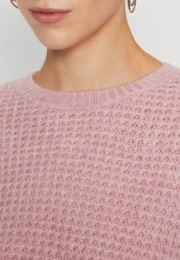 Ljusrosa stickad tröja med ett strukturerat mönster, rund halsringning och ribbade muddar. Det mjuka, fluffiga materialet ökar värmen och komforten.