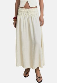 Falda maxi larga de color crema, con cintura elástica y fruncidos. Tela ligera con una textura suave y una silueta fluida.