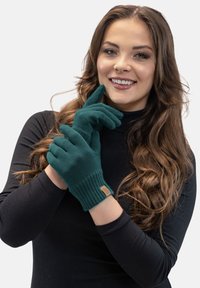 Guantes de lana de color verde azulado con puños acanalados, mostrados en un modelo. Presentan una pequeña etiqueta texturizada en la muñeca. Tejido suave y liso.