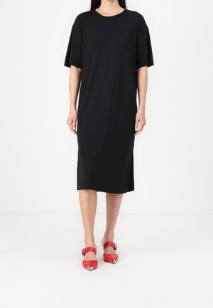 Donna che indossa un vestito midi nero largo con maniche corte e tacchi rossi a punta con cinturini alla caviglia, in piedi su un pavimento bianco.