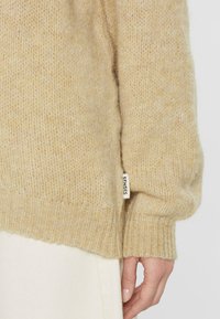 Beiger gestrickter Pullover mit strukturiertem Finish, gerippten Bündchen und einem kleinen Etikett am Saum. Weicher, gemütlicher Stoff, leicht oversized Schnitt.