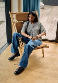 Cardigan gri cu mâneci scurte și nasturi, asortat cu blugi largi din denim albastru și pantofi negri lucioși, așezat pe un scaun din lemn.