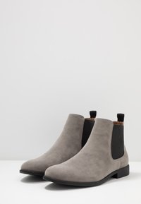 Botas de tornozelo em camurça cinza com painéis laterais elásticos pretos, solas planas e alças na parte de trás, apresentando um design elegante e aerodinâmico.
