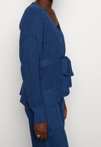 Blå stickad cardigan med v-ringning, långa ärmar och en knytdetalj i midjan. Texturerad tyg och en lagd nederkant.