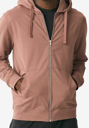 Haut en zip marron avec capuche à cordon de serrage, deux poches avant et poignets à côtes. Tissu doux au toucher lisse, superposé sur une chemise noire.