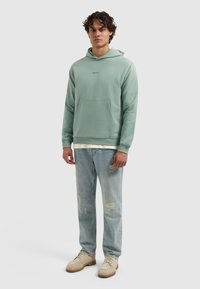 Man met krullend haar, gekleed in een mintgroene hoodie, licht verwaaide jeans en beige veterschoenen, staande tegen een effen achtergrond.