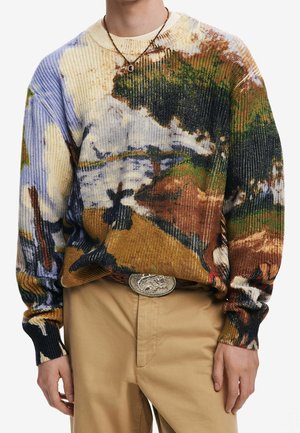Sweter na drutach z wielokolorowym abstrakcyjnym wzorem krajobrazu, w połączeniu z beżowymi spodniami i srebrną, owalną klamrą paska, noszony z prostym naszyjnikiem z zawieszką.